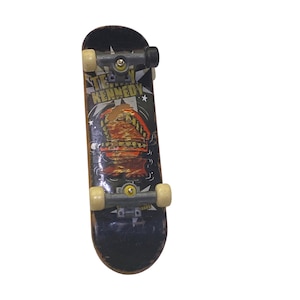 Könnte beinhalten: Ein schwarzes und braunes Fingerboard-Skateboard mit einem Motiv eines Cartoon-Monsters mit Zähnen und dem Text "Terry Kennedy". Das Skateboard hat schwarze Trucks und weiße Räder.