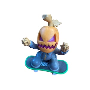Puede incluir: Una figura de color gris y naranja con forma de calabaza en una tabla de skate Tech Deck. La figura tiene una boca cosida y una cara morada.