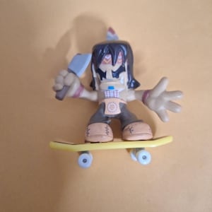 Könnte beinhalten: Eine Indianer-Figur auf einem gelben Skateboard. Die Figur hat einen Kopfschmuck, dunkle Haare und hält eine hellblaue Axt. Die Figur trägt eine braune Hose und beige Schuhe. Das Skateboard hat blaue Räder.