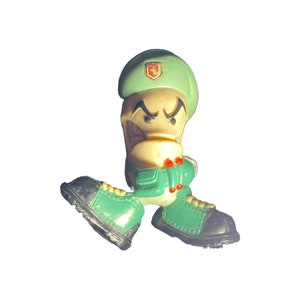 May include: Green and black toy figure with a green hat and a red and white symbol on the hat. The figure has a white face with black eyes and a black mouth. The figure is wearing green and black shoes.