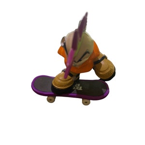 Puede incluir: Un monopatín de dedo morado y negro con una figura de juguete de una persona con ropa naranja y un mohawk rosa. La figura está de pie en el monopatín con un pie en la tabla y el otro pie en el aire.