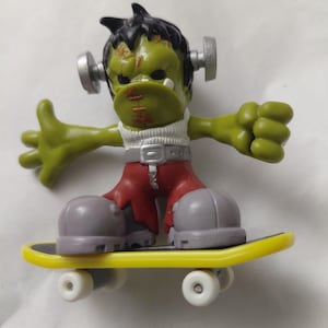 May include: A green Frankenstein-like figure with black hair, silver bolts, and red stitched mouth, wearing red pants and gray shoes, riding a yellow skateboard with white wheels. The figure has outstretched arms.