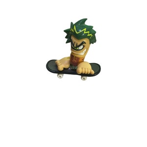 May include: A green and yellow fingerboard character with a mischievous expression rides a black fingerboard with white wheels.
