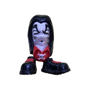 May include: A black and white figurine of a person with long black hair, wearing a red shirt with a white skull and crossbones design, and black boots.