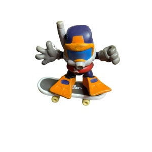 May include: A purple and orange toy figure wearing a diving helmet and flippers, riding a white and gray skateboard. The figure has a red star on its chest.