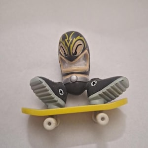 May include: A black and yellow figurine with a stylized face and a silver belt, wearing black shoes with white star accents, riding a yellow skateboard with white wheels. The figurine has a unique, cartoonish design.