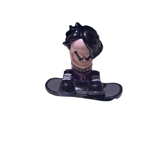 May include: A black and white figurine of a person with black hair wearing a purple and black outfit and riding a black skateboard.