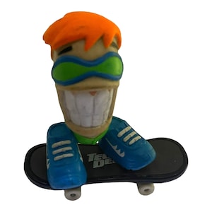 May include: A Tech Deck fingerboard with a smiling figure wearing green goggles and blue shoes. The figure is riding a black skateboard with white wheels and the words "Tech Deck" printed on the board.