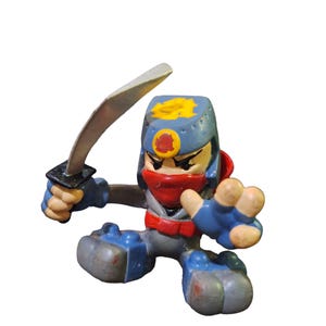 May include: A blue and gray action figure of a ninja holding a silver sword. The figure has a red bandana and a yellow symbol on its helmet.