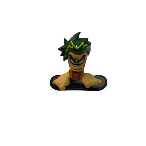 May include: A green-haired cartoon character with a red and yellow shirt is riding a black skateboard. The character has a mischievous expression on its face.