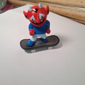 May include: A red devil figurine with blue clothing and white pants rides a small skateboard. The skateboard is gray with white wheels and the text "Tech Deck". The devil has white horns and a smiling face.