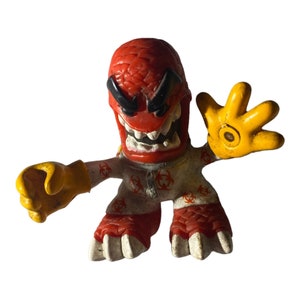 May include: Red and yellow plastic toy figure with a biohazard symbol pattern on its white clothing. The figure has a large, angry face with a wide mouth and sharp teeth. The figure has a raised hand with four fingers.