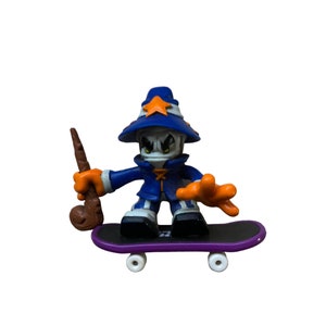 May include: A blue and orange fingerboard toy with a wizard character holding a brown staff. The wizard is wearing a blue robe and a blue hat with a star on top. The fingerboard is purple and black with white wheels.