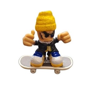 Puede incluir: Una figura de fingerboard con un gorro amarillo, una chaqueta negra y pantalones vaqueros azules montando un monopatín gris con ruedas negras.
