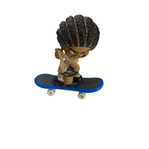 May include: A small, brown-skinned figurine of a person with black braided hair, wearing a white shirt and black pants, is holding a skateboard with blue wheels and a blue deck. The figurine is performing a trick on the skateboard.