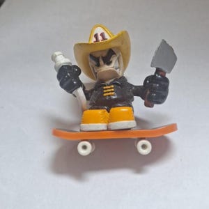 Puede incluir: Una pequeña figura de un pato con un sombrero de vaquero amarillo con el número 11, una chaqueta negra y botas naranjas. El pato sostiene un objeto blanco en una mano y un hacha pequeña en la otra. Está montado en una patineta naranja.