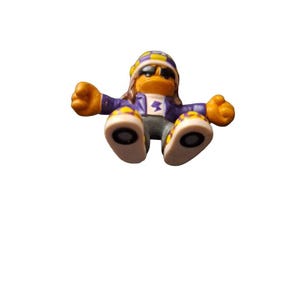 Puede incluir: Una pequeña figura de plástico de una persona con un sombrero y gafas de sol. La figura es de color morado y amarillo, tiene un rayo en el pecho y está sentada con las piernas cruzadas.