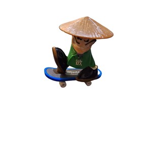 May include: A green and brown figurine of a person wearing a conical hat and holding a skateboard. The figurine is on a blue skateboard with black wheels. The figurine has the word "欲" written on its chest.