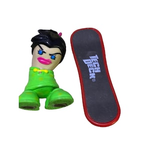 May include: A green and black Tech Deck fingerboard with a female figure with black hair and blue eyes. The figure is wearing a green shirt and pants.