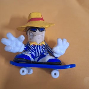Puede incluir: Una pequeña figura novedosa de un personaje que lleva un sombrero amarillo con una banda roja, gafas de sol y un traje a rayas azul y blanco. La figura tiene guantes blancos y monta en un monopatín azul con ruedas blancas.