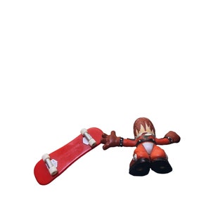 Puede incluir: Un monopatín rojo para fingerboard con un camión y ruedas plateadas. Una figura roja y negra de una persona con el pelo de punta sostiene el monopatín.