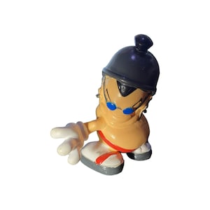 May include: A small figurine of a cartoon character with tan skin, wearing a black helmet, blue sunglasses, and white gloves. The figure has a red stripe on the bottom of the feet. The figurine is posed with one arm extended.