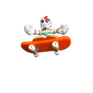 Può includere: Un fingerboard bianco e arancione con una figura di un personaggio con un vestito bianco e blu e un fulmine rosso sulla testa. La figura è in una posa da skateboard con le braccia distese.