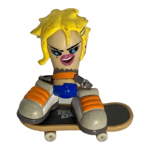 May include: A yellow-haired figure skater with a blue and orange outfit is riding a Tech Deck skateboard. The figure has a painted face with blue eyes and a pink mouth.