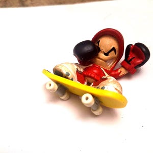 May include: A small figurine of a boxer wearing a red hoodie and boxing gloves, riding a yellow skateboard. The boxer has a black eye and a mischievous grin. The skateboard has white wheels.