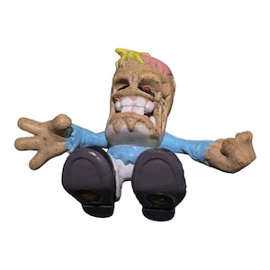 May include: A small, brown, and white figurine of a cartoon character with a cracked head, a blue shirt, and purple shoes. The character has a wide, toothy grin and a red eye.