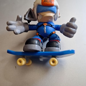 Puede incluir: Una figura de juguete azul y blanca que lleva un casco con "ACE" impreso, un mono azul y guantes grises, montada en una patineta azul con ruedas amarillas. La figura está haciendo una señal de pulgar hacia arriba.