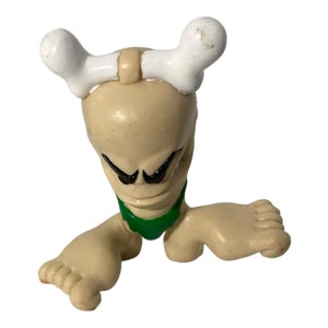 May include: A small, beige figurine with a white bone headband and green bikini bottom. The figurine has a cartoonish alien-like face with black eyes and a wide mouth.