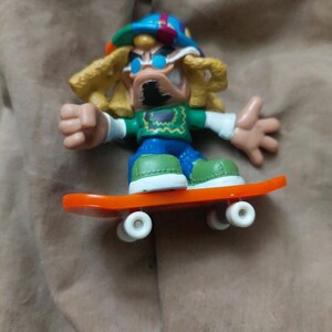 May include: A small, colorful skateboard toy featuring a cartoon character with sunglasses, a hat, and blonde hair. The character is wearing a green shirt, blue pants, and green shoes. The skateboard is orange with white wheels.