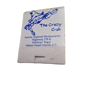 May include: A white matchbook with blue text and a cartoon crab illustration. The text reads "The Crazy Crab Family Seafood Restaurants Highway 278 & Harbour Town Hilton Head Island, S.C."