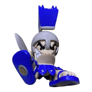 May include: A small, cartoonish figurine of a gladiator with a white helmet, blue accents, and a sword. The figurine has a menacing grin and is holding a blue shield. The gladiator is wearing blue sandals.