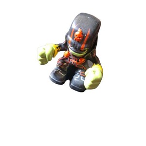 May include: Black and orange action figure with a skull design on the head. The figure has green hands and is wearing black boots.