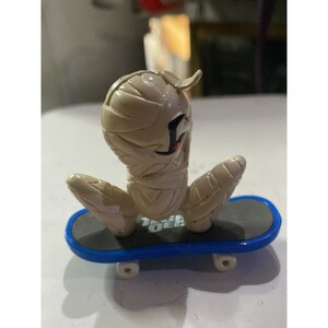 May include: A tan mummy figure riding a blue and gray fingerboard skateboard. The mummy has a black eye and a red mouth.