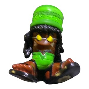 Può includere: Figurina verde e nera di una persona che indossa un cappello verde con la scritta "RISKY". La figurina ha un viso marrone con occhi gialli e una camicia verde. La figurina è seduta con le gambe incrociate.