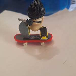 Peut inclure: Un jouet de skateboard miniature avec un personnage souriant, une coiffure noire, des genouillères et un microphone. Le skateboard a une planche rouge, des roues blanches et un grip gris.