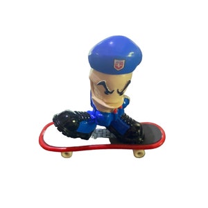 May include: A blue and black fingerboard toy with a red skateboard and a character wearing a blue beret with a white cross on it.