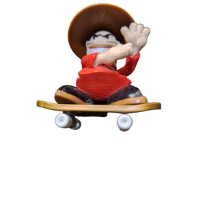 May include: A red and black figurine of a person wearing a brown hat and riding a skateboard. The figurine is in a sitting position with one hand raised.