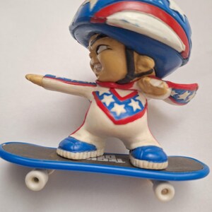 May include: A figurine of a person skateboarding, wearing a white jumpsuit with red and blue star accents, a blue helmet with white stars, and blue shoes. The skateboard is black with blue wheels.