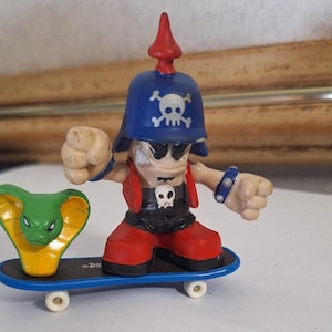 Puede incluir: Una figura de juguete de un pirata en una patineta. El pirata lleva un casco azul con una calavera, un chaleco rojo y botas rojas. Una cobra verde y amarilla está al lado del pirata. La patineta es azul con ruedas blancas.