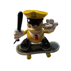 May include: A yellow and white figurine of a police officer riding a skateboard. The officer wears a yellow hat with "COP" on it, a white shirt, and a red tie. He holds a black baton and a donut. The skateboard has "TECH DECK" printed on it.