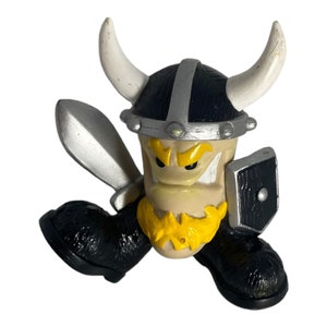 May include: A black and silver plastic toy figure of a Viking warrior wearing a horned helmet, holding a sword and shield. The figure has a yellow beard and angry facial expression.