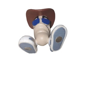 Puede incluir: Una pequeña figura de plástico de una persona que lleva un sombrero de vaquero marrón, gafas de sol azules y zapatos blancos con suelas azules. La figura está boca abajo.