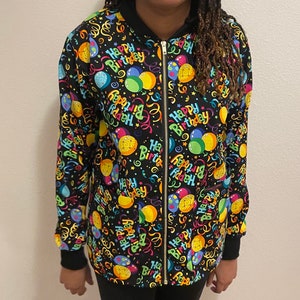 May include: Black zip-up jacket featuring a vibrant "Happy Birthday" design. The pattern includes balloons, confetti, and stars in multiple colours. The jacket has a black collar, cuffs, and a gold zip.