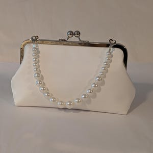 May include: A white satin clutch with a silver clasp and a pearl beaded handle. The clutch has a rectangular shape with rounded edges. The pearl handle is attached to the clasp with small silver connectors. The bag is designed for formal events.