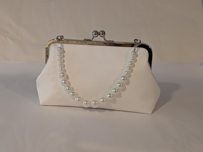 May include: A white satin clutch with a silver clasp and a pearl beaded handle. The clutch has a rectangular shape with rounded edges. The pearl handle is attached to the clasp with small silver connectors. The bag is designed for formal events.