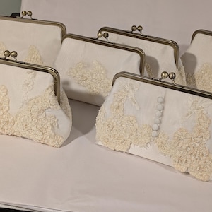 May include: Several off-white clutch purses with metal clasps are displayed. The purses are adorned with delicate, cream-colored lace detailing. Some purses have a row of small, white buttons. The purses are arranged on a white surface.